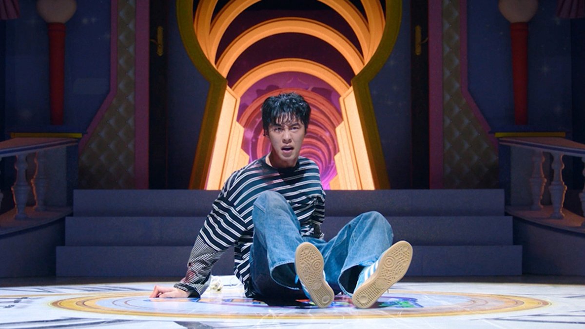 Lee Jae-wook, wearing a striped shirt and jeans, sits on the floor with a startled expression in front of a colorful, theatrical set with a glowing archway, capturing the whimsical and surreal tone of his new Korean series "A Hell of a Job." The image visually emphasizes his dramatic reaction, aligning with the post's playful question about taking on this otherworldly freelance gig premiering October 30.