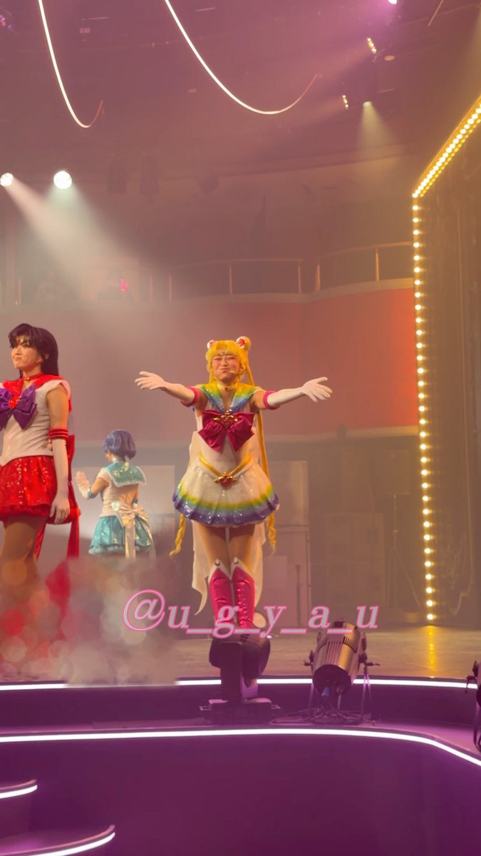 A performer in a Sailor Moon costume with blonde pigtails strikes a pose on stage with arms outstretched, with a watermark '@u_g_y_a_u' visible.