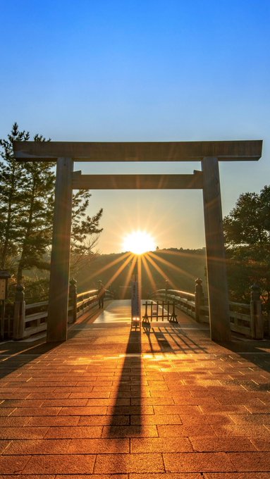 A torii gate at Ise Grand Shrine stands silhouetted against the rising sun, casting long shadows across the stone path in a serene, golden-hued scene. The image perfectly matches the post's promotion of a fortune-bringing waiting screen, capturing the auspicious sunrise through the iconic shrine entrance.
