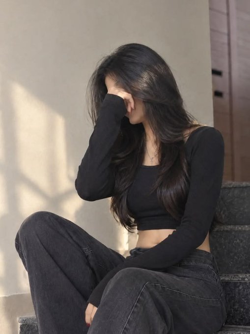 A woman with long dark hair wearing a black long-sleeved crop top and dark jeans sits on steps with her hand covering her face.