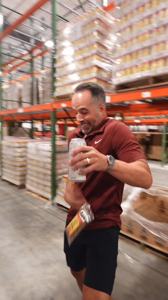 A man in a maroon polo shirt and dark shorts walking through a warehouse-style store while holding a can and a packaged item.