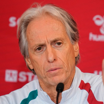 A close-up portrait of an older man with gray hair wearing a white shirt with green and red accents, seated in front of a red backdrop with partial logos visible.