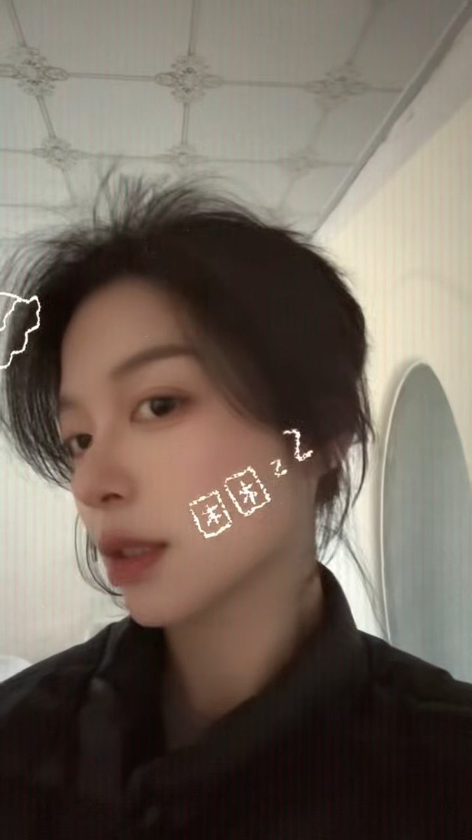 A selfie of a young woman with dark hair in a black shirt, with a drawn "Zzz" and "困" (meaning sleepy) on her cheek, suggesting she's tired.