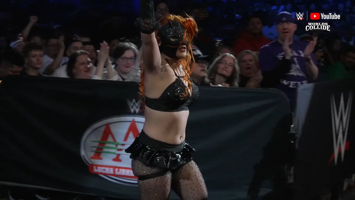 A WWE wrestler with orange hair, wearing a black mask and revealing outfit, poses in the ring with her arm raised as the crowd cheers behind her; logos for WWE and Worlds Collide are visible.