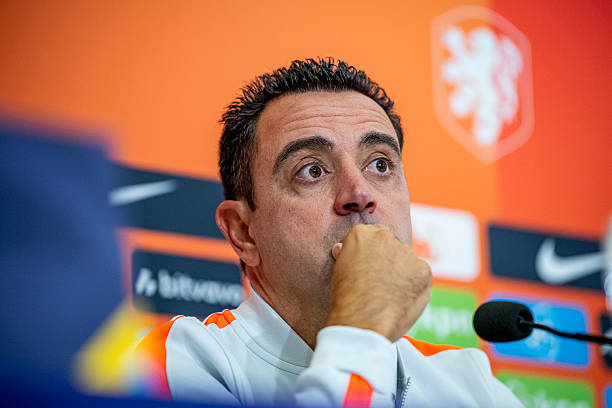 A man in an orange and white jacket appears thoughtful at a press conference, with a Dutch lion logo and Nike branding visible in the background.