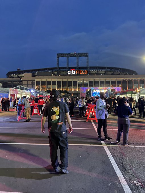 An evening view of Citi Field stadium with a crowd of people gathered outside in a parking lot area.