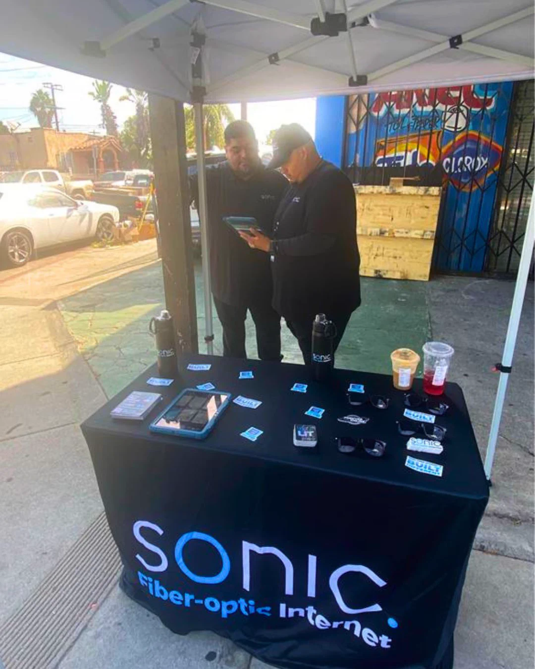 Outdoor Sonic Fiber Internet booth under a canopy tent, with Field Sales team members Raul and Johnny reviewing a tablet behind a table displaying Sonic flyers, sunglasses, and stickers. 