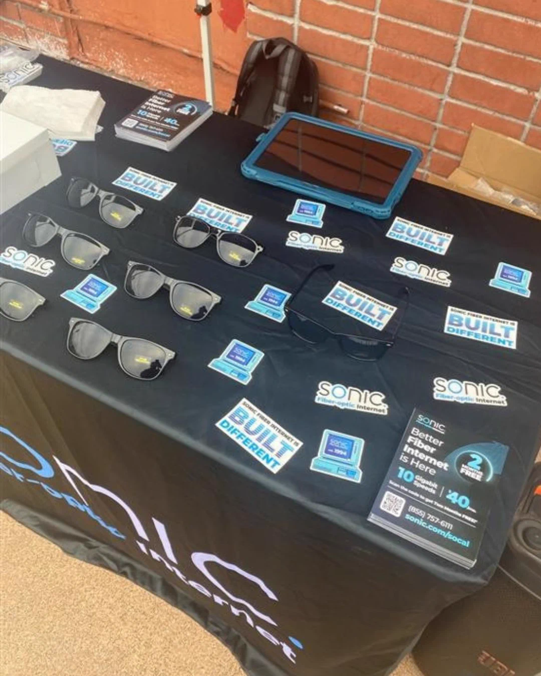 Table covered with Sonic Fiber Internet merch, featuring Sonic sunglasses, stickers, and flyers, set up outdoors in front of a brick wall. A tablet rests on the table as well.  