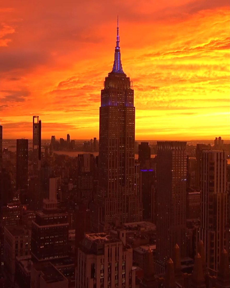 An aerial cityscape view of the Empire State Building illuminated against a dramatic orange and red sunset sky.