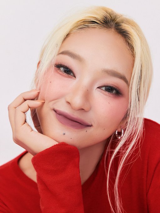 A close-up portrait of a smiling woman with blonde hair and pink-tinged strands, wearing a red sweater and red hoop earrings, resting her chin on her hand.