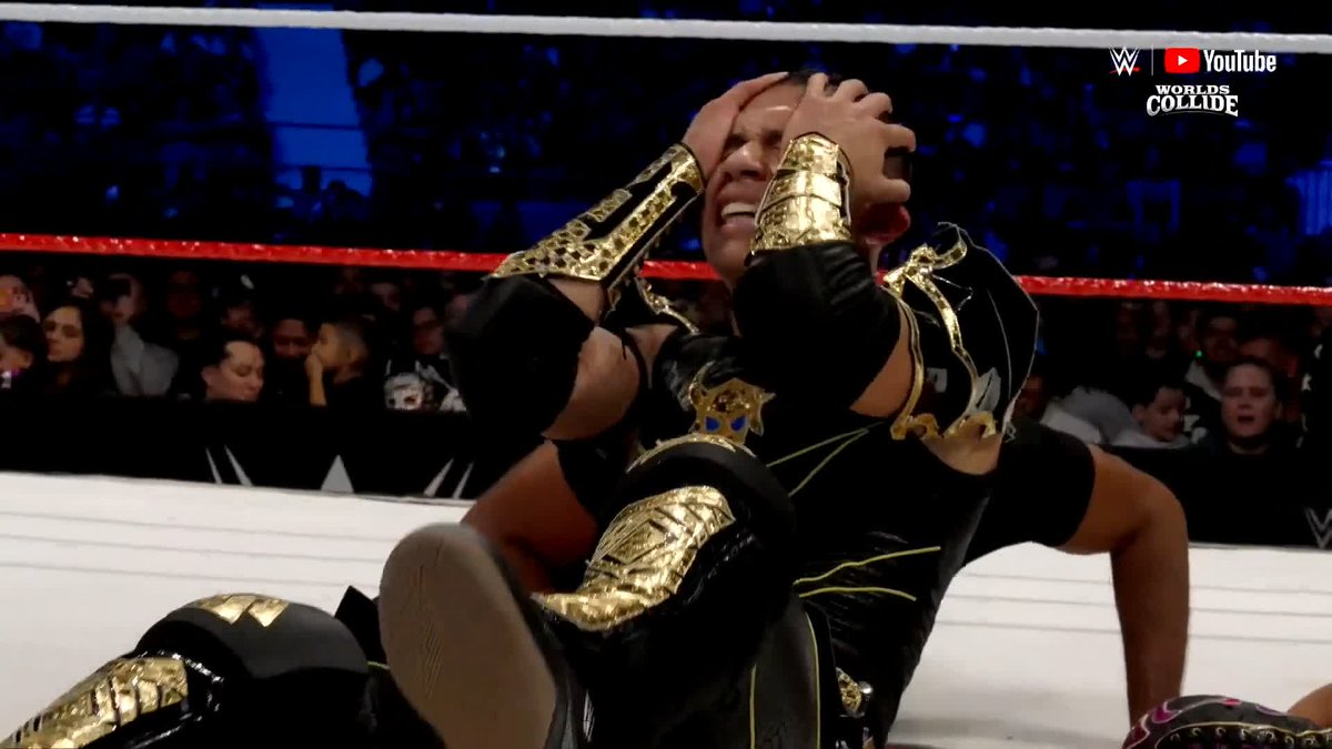 A WWE wrestler in black and gold attire holds his head in apparent pain or disbelief on the mat during the Worlds Collide YouTube broadcast.