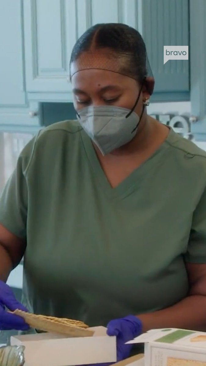 A woman with her hair pulled back, wearing a gray face mask, olive green scrubs, and purple gloves, looks down while handling what appears to be a cracker over a box in a kitchen with light blue cabinets. A speech bubble graphic in the upper right reads "bravo," tying the scene to the post about Mary from #RHOSLC navigating grief.