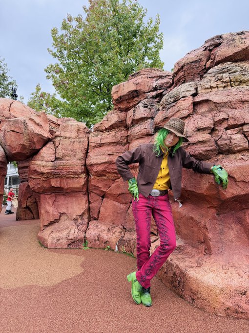 A person cosplaying as Gyro Zeppeli from JoJo's Bizarre Adventure, wearing green hair, a brown hat, brown jacket, yellow shirt, magenta pants, and green gloves, posing against a red rock formation.