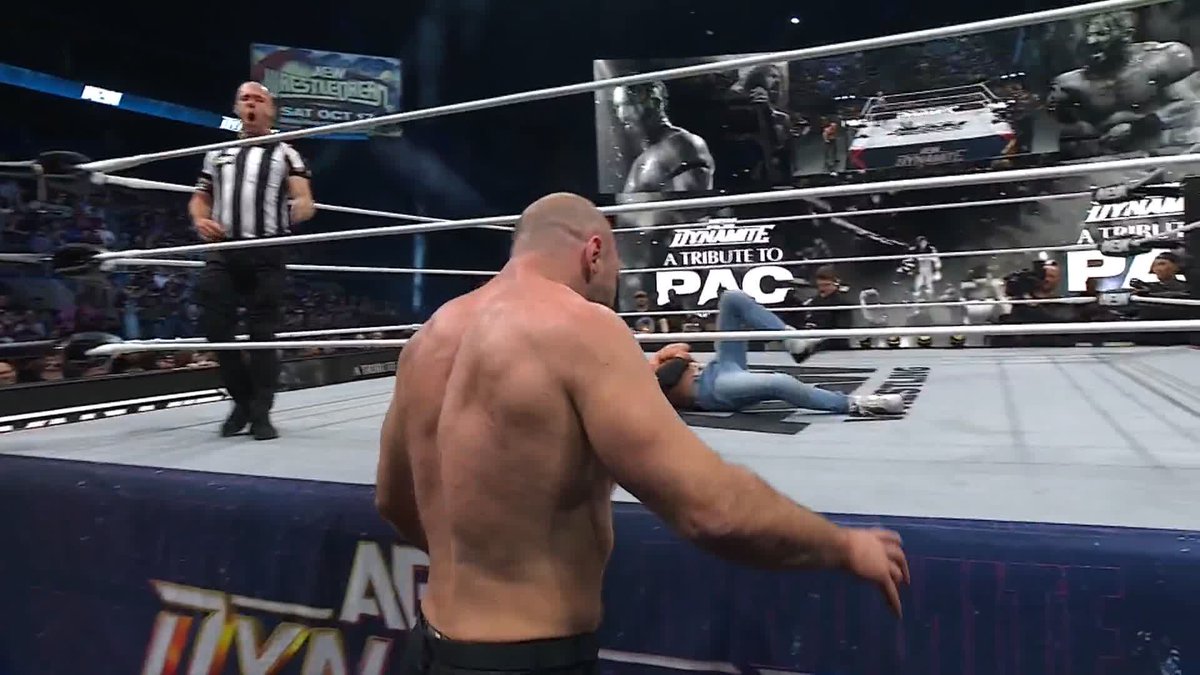 A wrestler stands outside the ring watching his opponent, wearing blue jeans, lie on the mat, with referee in the ring nearby. The arena screens display "DYNAMITE A TRIBUTE TO PAC" along with "AEW WRESTLEFEST SAT OCT 15," connecting to the post about Orange Cassidy's tactics on the special AEW Dynamite episode.