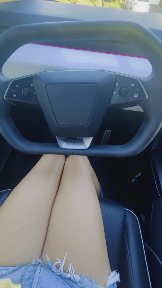 A first-person view from the driver's seat of a car, showing bare legs and denim shorts beneath a yoke-style steering wheel with control buttons, looking out toward a road with other vehicles.