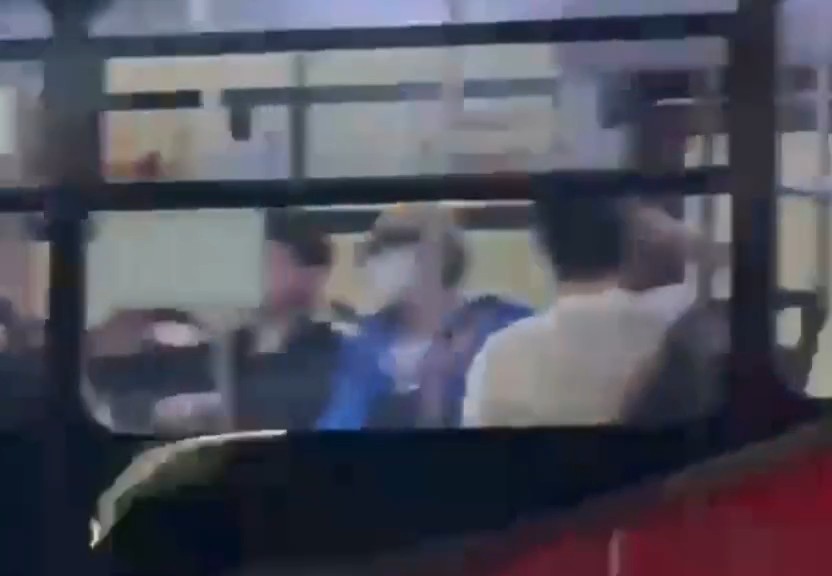 Blurry photo through a bus window showing people seated inside, including one in a blue shirt and another in a light-colored jacket.