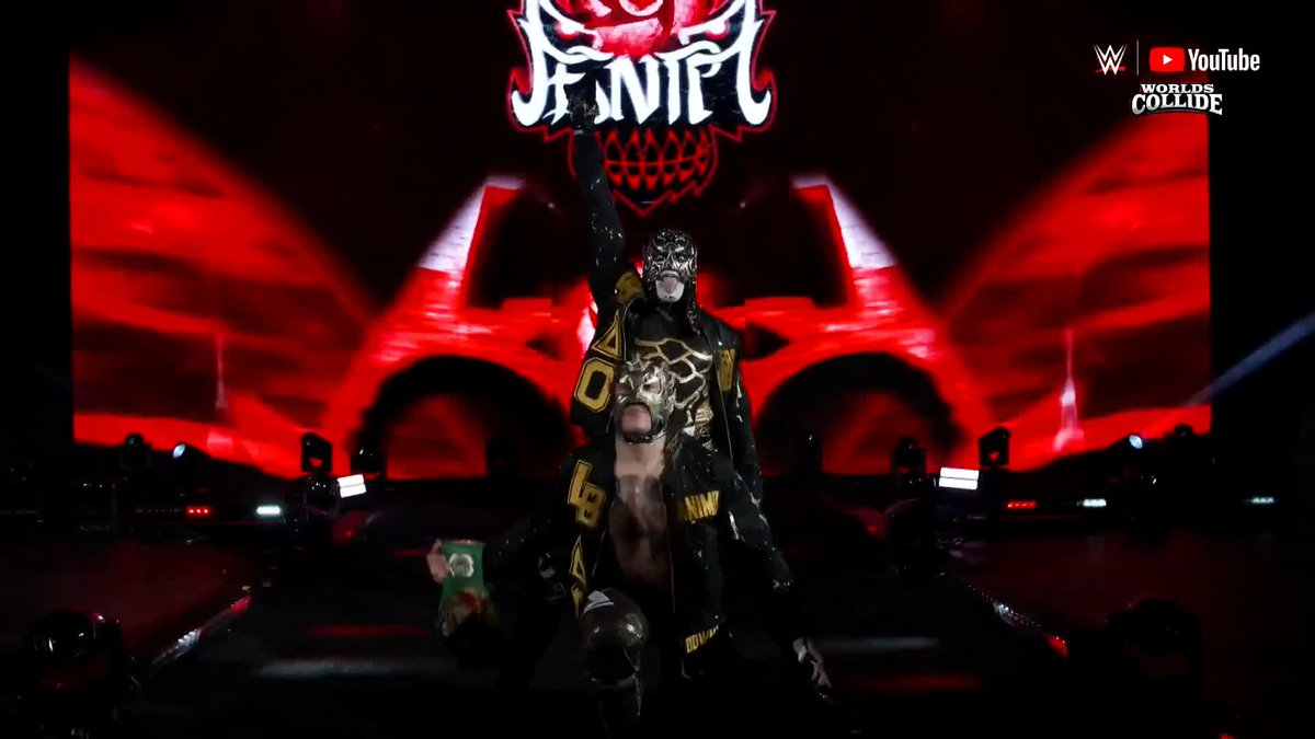 A masked wrestler wearing black and gold gear with "Lucha" lettering makes a dramatic entrance, raising his fist in front of a large screen displaying his name and a skull graphic, while a "WWE Worlds Collide" logo with YouTube branding appears in the top right corner.