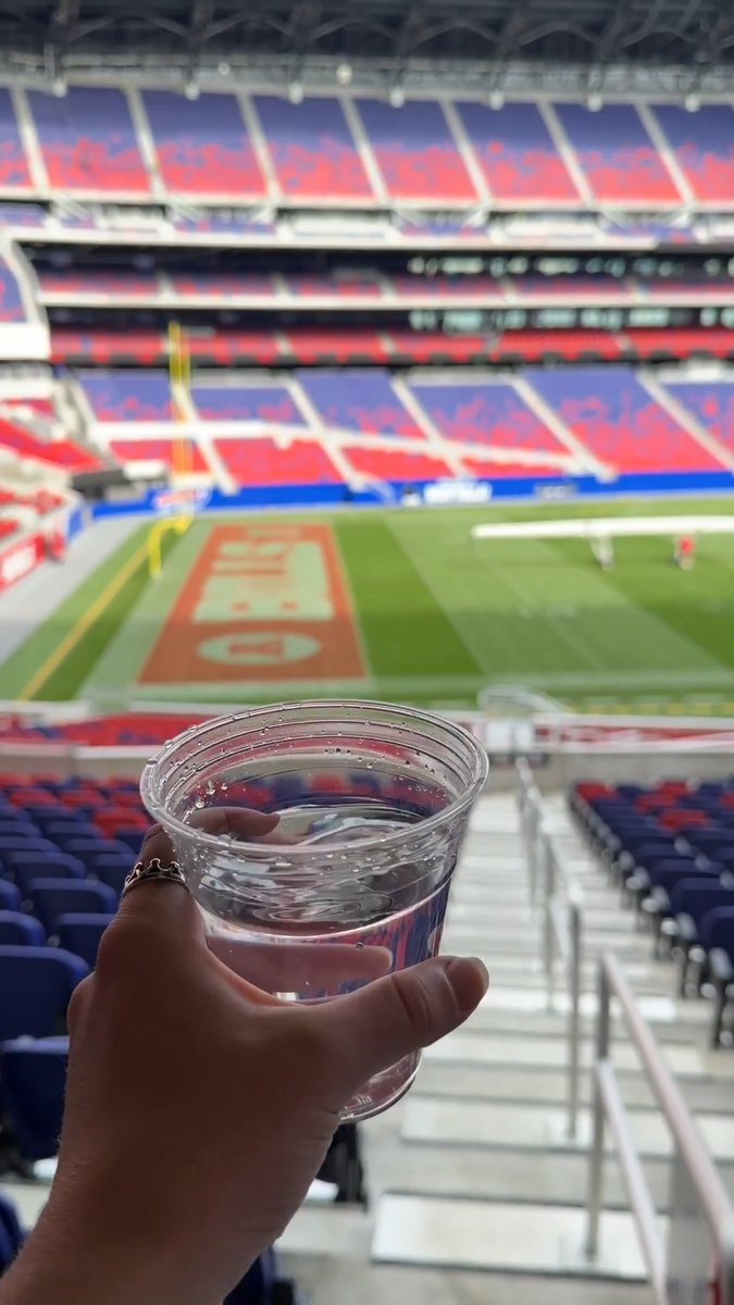 A hand raises a clear water cup with a view of the empty Highmark Stadium field, showing the "BILLS" endzone logo, before kickoff. The image illustrates the post about complimentary water cup locations for Bills Mafia fans on game day.