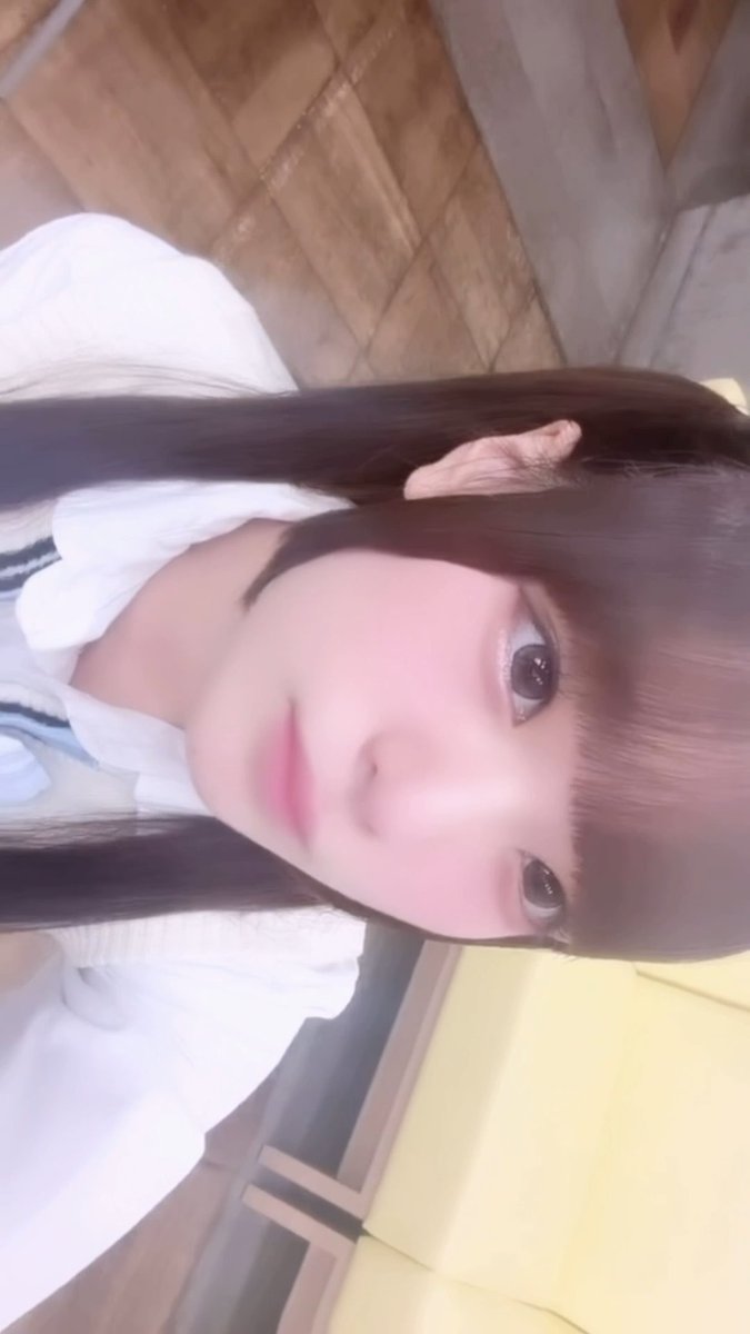 A sideways-oriented selfie of a young woman with long dark hair, soft makeup, and pink lips, wearing a white top with a bow detail.