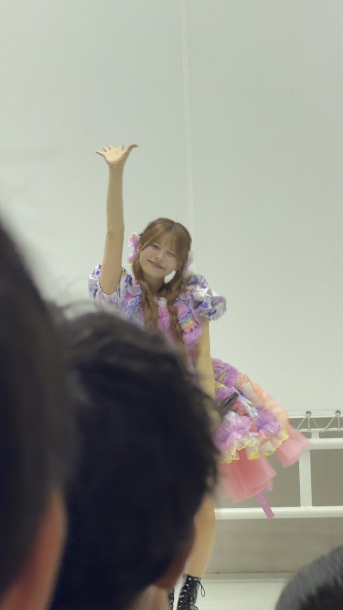 A performer in a pink and purple frilly dress waves to the audience from a stage.