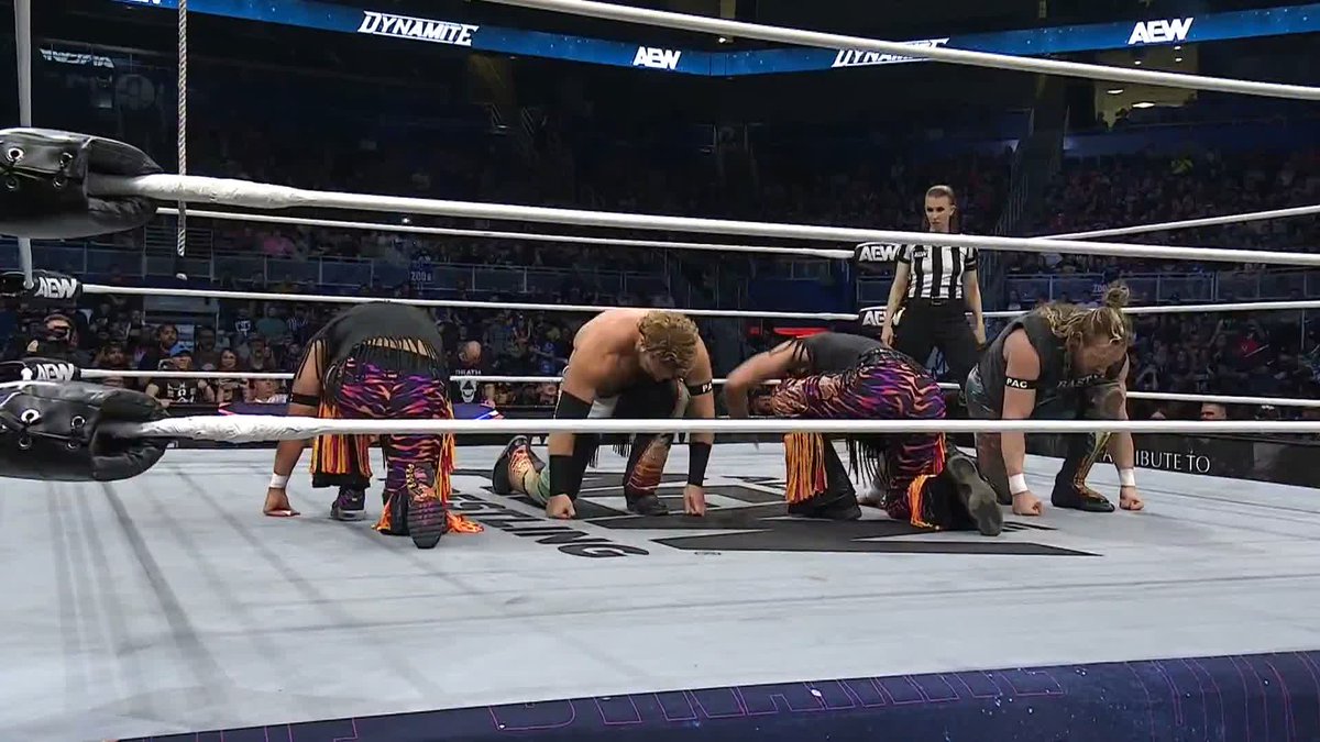 Four AEW wrestlers, including PAC, prepare to launch a quadruple topes suicida simultaneously in the ring at the "Tribute to PAC" event, while the referee stands ready to observe. The ring ropes and surrounding signage display "AEW Dynamite" branding, highlighting the chaotic moment from this tribute episode.