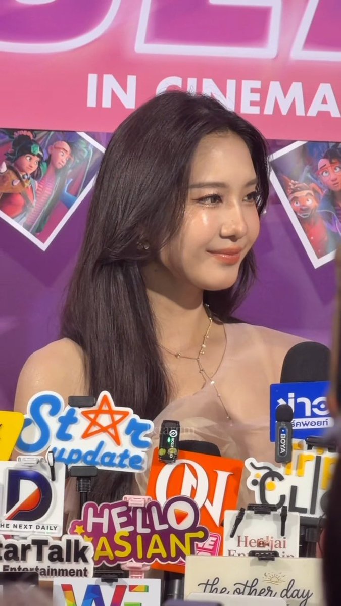 A woman with long dark hair wearing a sheer beige top and pearl necklace speaks at a media event surrounded by microphones from outlets like Star Update, Hello Asian, Star Talk, and The Other Day, with a movie promotional poster behind her.