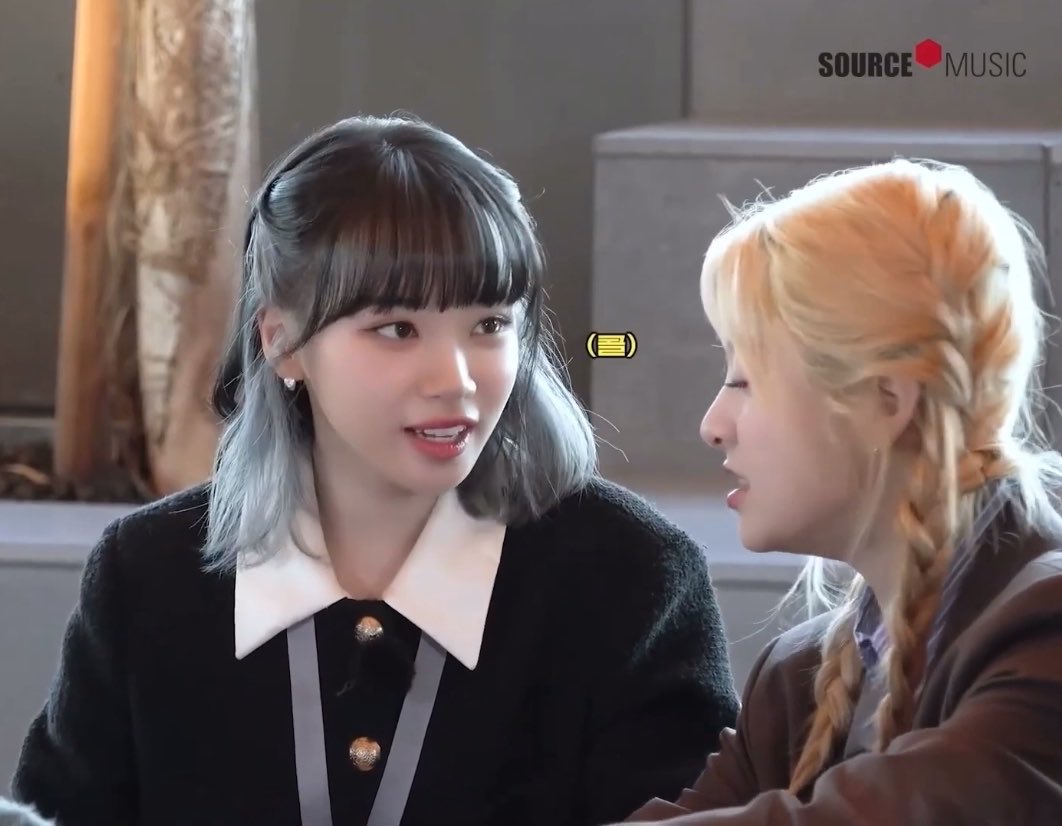 Two women converse in an indoor setting with a "SOURCE MUSIC" logo in the corner; one has dark hair with silver highlights and a black collared top, while the other has blonde braided hair.