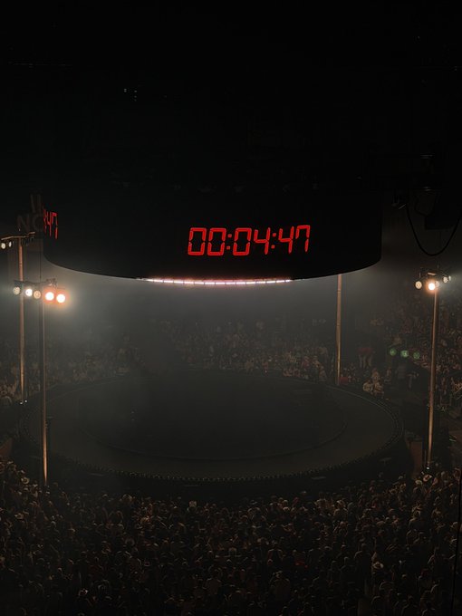 A circular countdown timer displaying '00:04:47' in red digital numbers suspended above a circular arena or stage, surrounded by a large crowd in dim lighting.