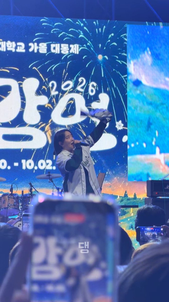 A performer in a white jersey sings into a microphone on stage during a concert, with a large LED screen behind displaying "2026" and Korean text along with fireworks graphics, while audience members hold up phones.