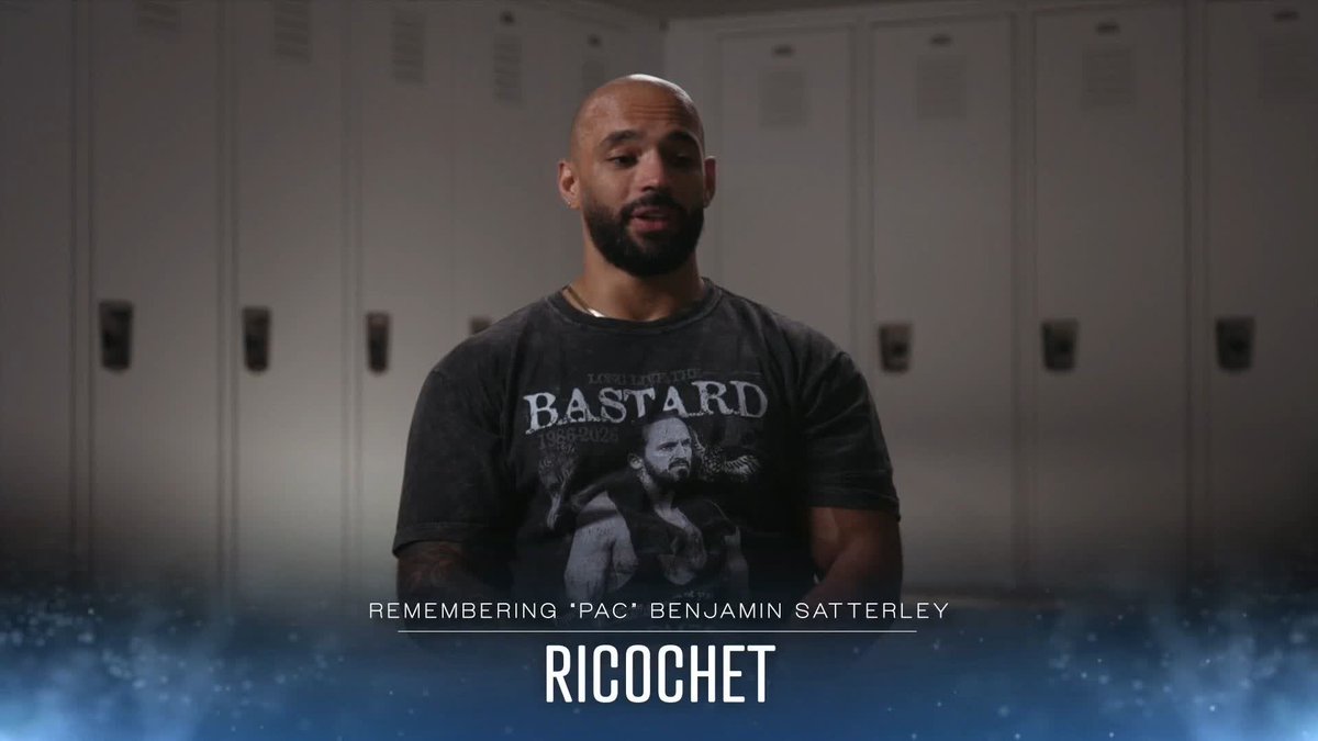 A bearded man stands in what looks like a wrestling locker room, wearing a "Long Live the Bastard" t-shirt, with on-screen text reading "REMEMBERING 'PAC' BENJAMIN SATTERLEY" and "RICOCHET" identifying him as wrestler Ricochet paying tribute to his fallen colleague.