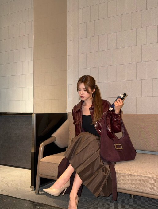 A woman in a burgundy leather jacket and brown pleated skirt sits on a beige sofa, holding a smartphone and a large matching handbag.