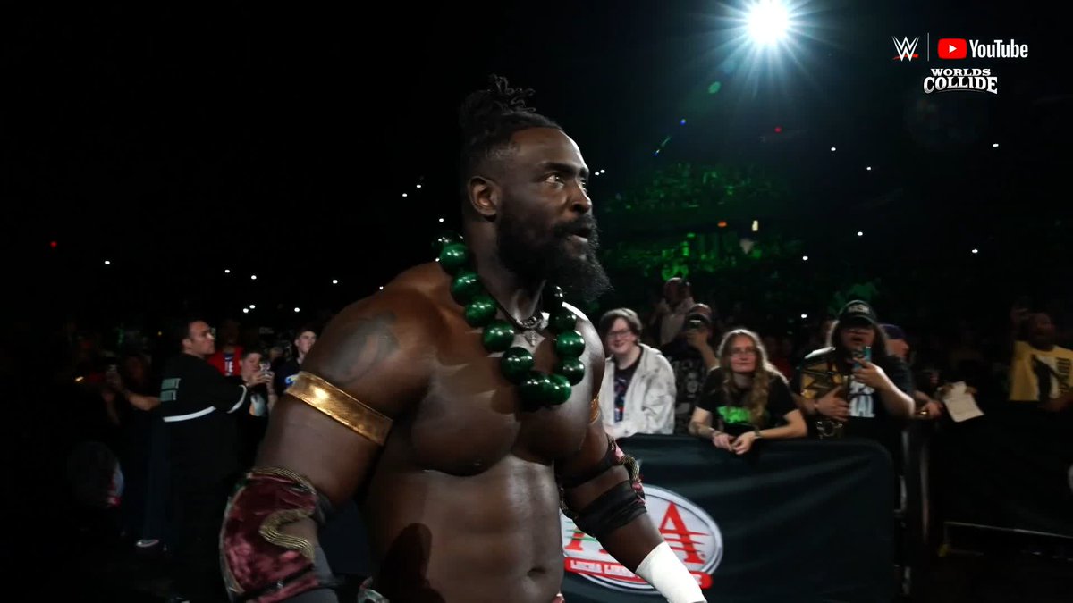 A bearded wrestler wearing a large green bead necklace stands in the arena during a WWE Worlds Collide event streamed on YouTube, as fans cheer behind the barricade. The "OOOOOBA!" chant in the post reflects the crowd's enthusiastic reaction to this charismatic performer's entrance.