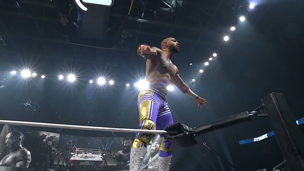 A wrestler stands triumphantly atop the ring ropes in purple and gold attire, silhouetted dramatically against bright arena spotlights. The visible "Dynamite" branding on the apron connects to the caption about #AEWDynamite, suggesting this is a dramatic moment from the tribute episode to PAC.