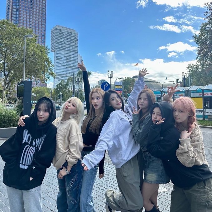 A group of seven young women pose together outdoors on a city street with skyscrapers in the background.