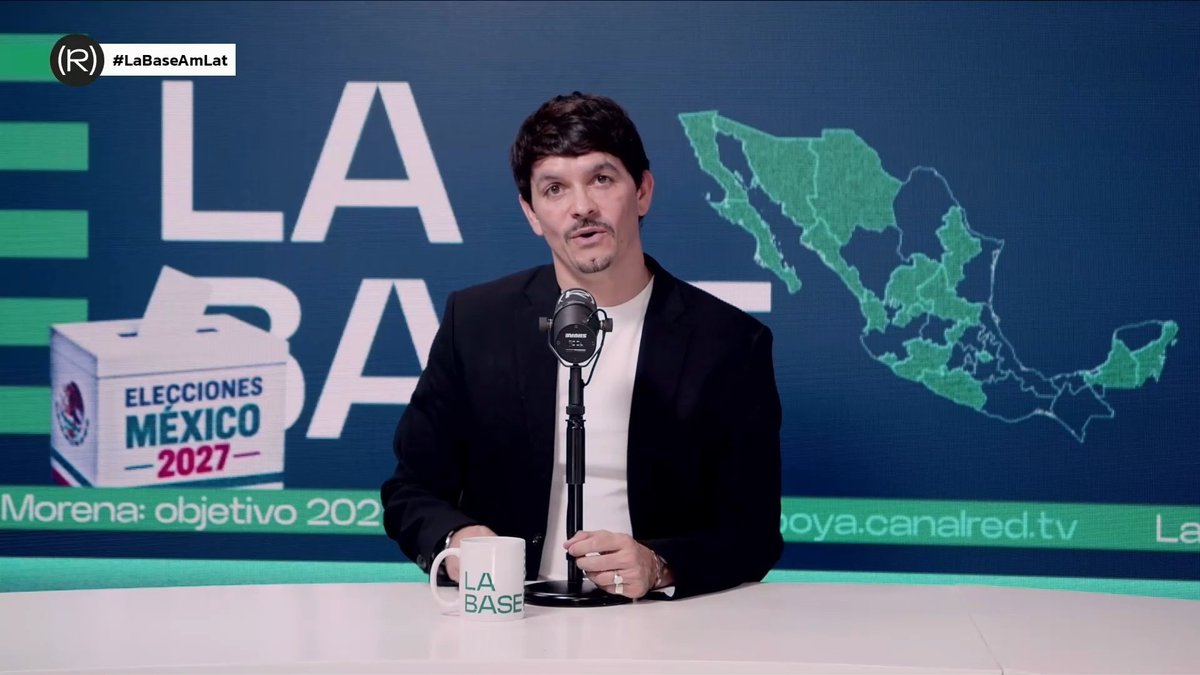 A man in a dark blazer and white t-shirt sits at a desk with a microphone and a 'LA BASE' mug, in front of a screen displaying 'LA BASE', 'Elecciones México 2027', and a map of Mexico.