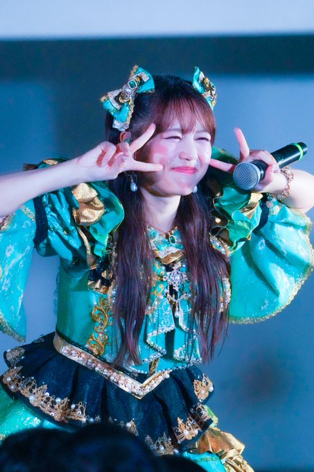 A performer on stage, wearing an elaborate teal and gold outfit with cat-ear hair accessories, winking and smiling brightly at the audience.
