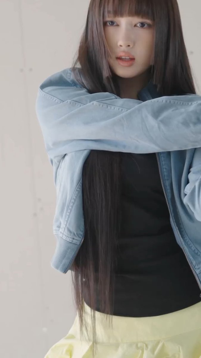 Lisa from BLACKPINK poses for a photo wearing a light blue jacket draped over her shoulders, a black top, and a pale yellow skirt against a concrete wall.