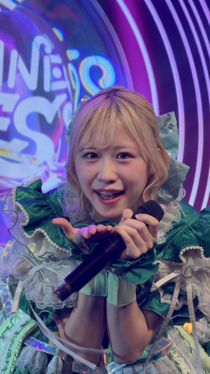 A person with short pinkish-blonde hair in an elaborate green and white ruffled costume holds a microphone close to their mouth on stage, with "Hello" partially visible on the colorful backdrop behind them, relating to the concert or performance context of the "@nowstorage_umya" post.