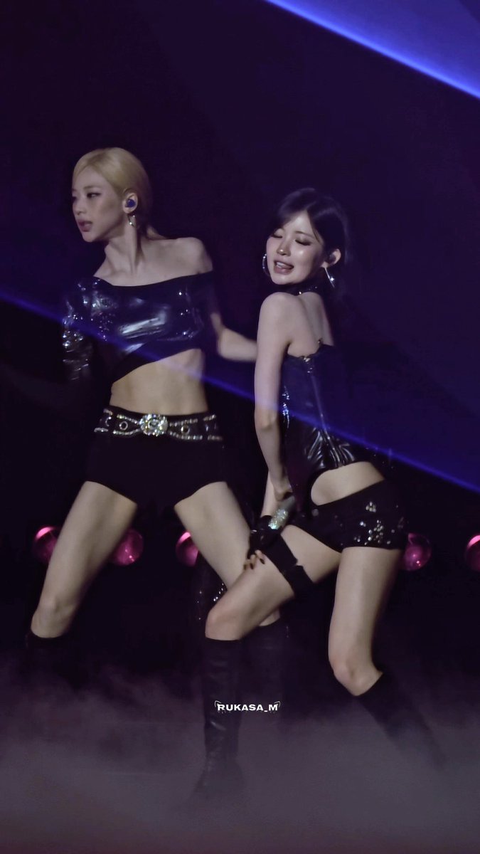 Two female K-pop performers in dark, sparkly stage outfits perform a dance routine under purple stage lighting, with the watermark 'RUKASA_M' at the bottom.