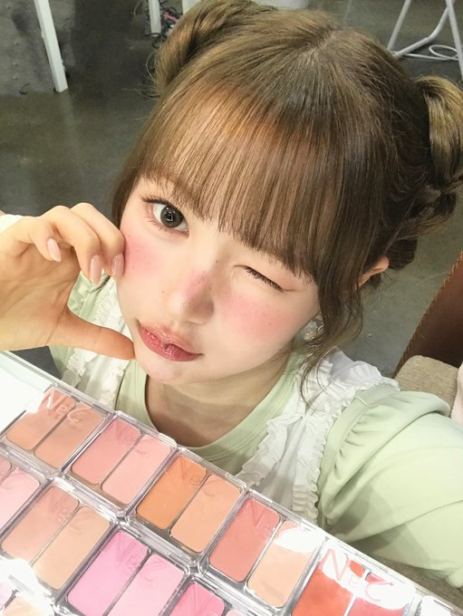 A young woman with brown hair in twin buns poses playfully while winking, holding up her hand near her rosy cheeks, with a colorful multi-pan blush palette open in the foreground.