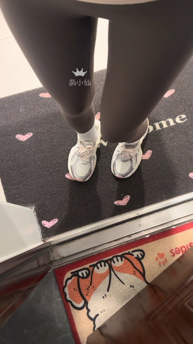 A photograph showing a person's legs in black leggings and white/silver sneakers standing on a black doormat decorated with pink hearts, with another illustrated mat visible at the bottom.