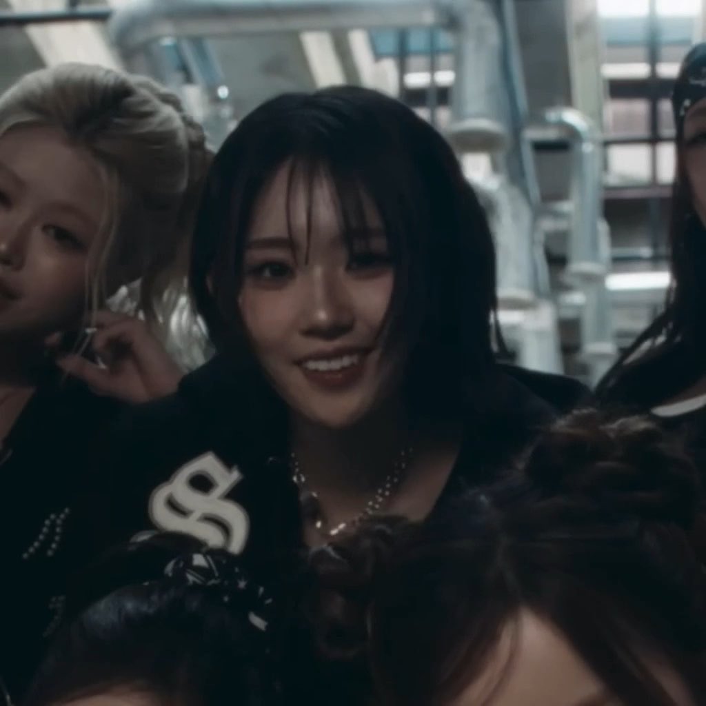 A woman with dark hair and bangs smiles at the camera while wearing a black jacket with white lettering, standing in a futuristic-looking setting alongside two other women, one with blonde hair in pigtails.