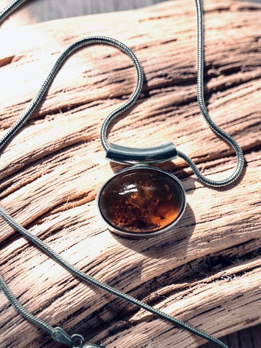 A round, dark stone pendant necklace with an amber/orange interior rests on a weathered wooden surface, connected to a silver snake chain.