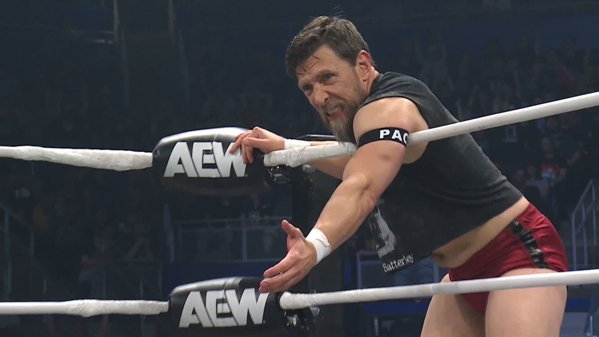 Bryan Danielson wrestles in the ring at an AEW Dynamite event, wearing a black sleeveless shirt, red trunks, and a "PAC" armband while leaning over the ropes. The ring posts display prominent "AEW" branding, tying the scene directly to the post's enthusiastic commentary about his performance.