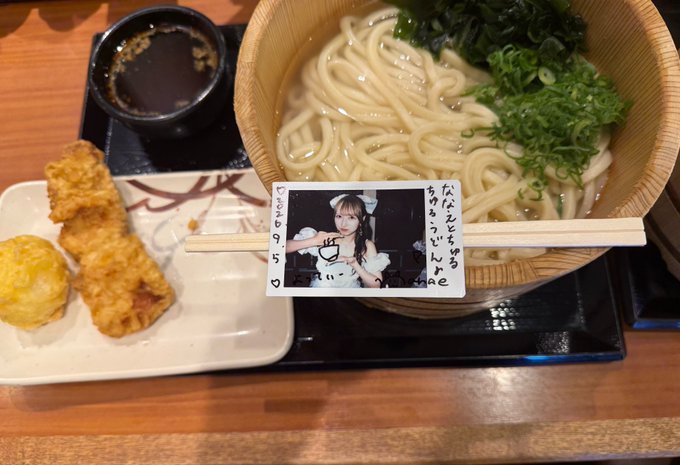 A Japanese meal set features a bowl of udon noodles with green onions, a side of tempura, dipping sauce, and a personalized photo card with handwritten Japanese text.