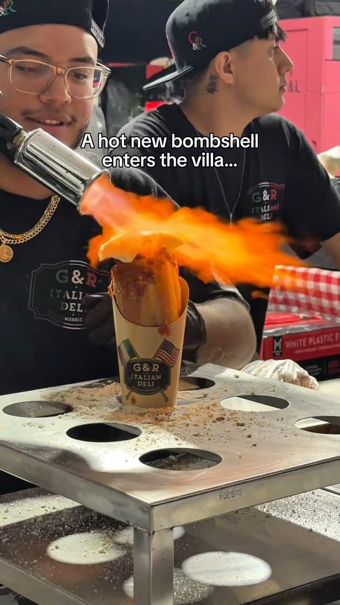 A meme image showing two men at G&R Italian Deli preparing food with a blowtorch creating fire on a sandwich, with caption 'A hot new bombshell enters the villa...'.