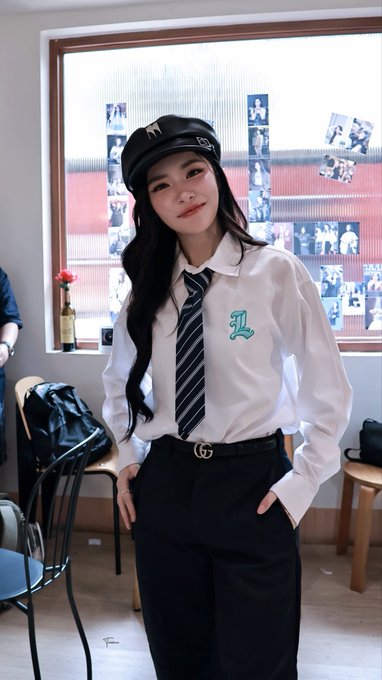 A young woman poses in a preppy schoolgirl-style outfit featuring a black cap with a lion emblem, a white collared shirt with a teal 'L' logo, a striped tie, and black trousers with a Gucci belt.