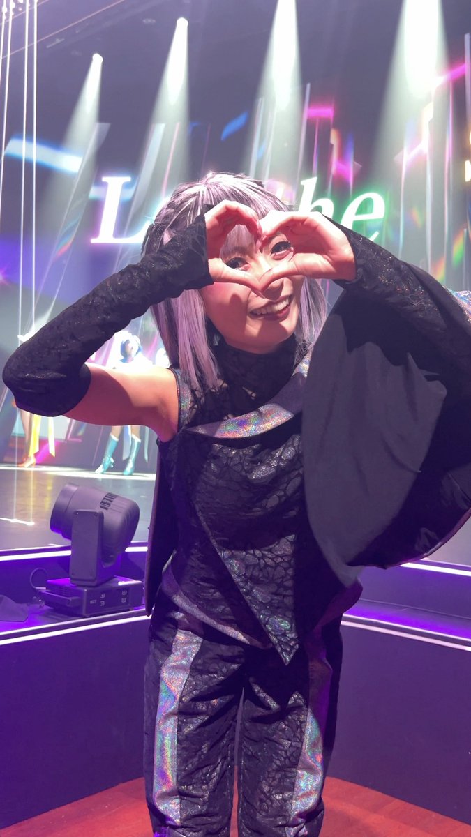 A performer on stage with light purple hair wearing a black, shiny, textured outfit and long black gloves, making a heart shape with her hands over her eyes, with stage lights and a partial 'Like' sign visible in the background.