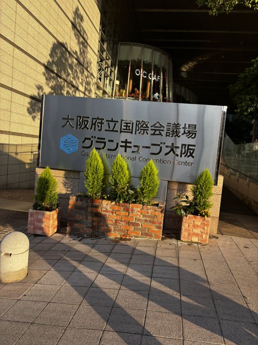 The exterior entrance sign of the Osaka International Convention Center (Osaka-kenritsu Kokusai Kaigijō), also labeled 'グランキューブ大阪' (Grand Cube Osaka), flanked by brick planters with green shrubs.