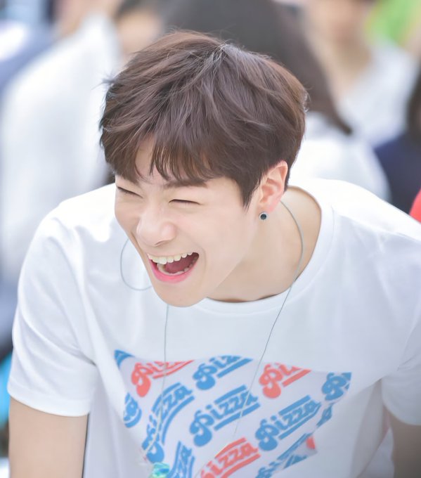 A young man with brown hair laughs heartily, eyes closed and mouth wide open, wearing a white t-shirt with colorful 'Pizza' text and an earphone in his left ear.